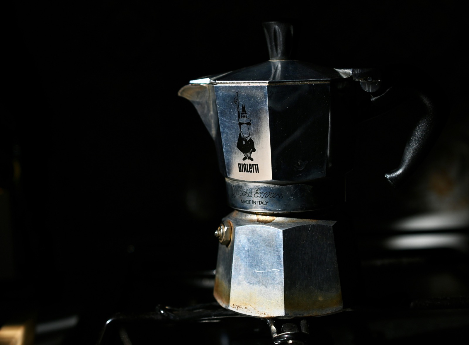 A well-used Bialetti Moka Express on a stove, its aluminum body lit against a dark background